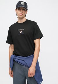 Czarny bawełniany t-shirt z haftowanym logo „PENFIELD” i grafiką. Jasnoniebieska bluza zawiązana w pasie, szare spodnie o luźnym kroju.