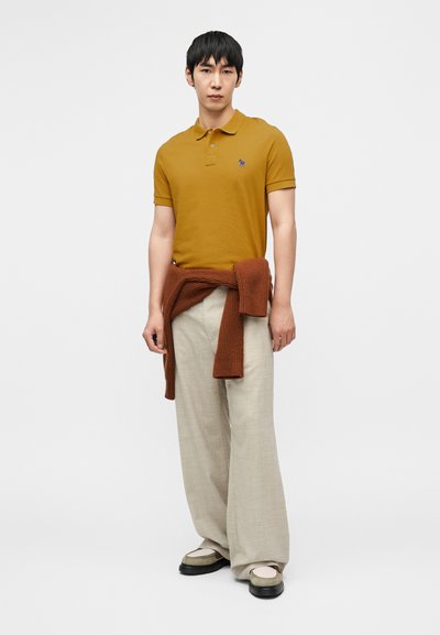 Μουσταρδί πόλο πουκάμισο με κοντά μανίκια και λογότυπο, συνδυασμένο με ανοιχτό μπεζ παντελόνια με φαρδιά πόδια και καφέ πλεκτό πουλόβερ δεμένο γύρω από τη μέση.