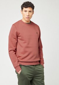 Uomo con capelli corti e ricci, che indossa un felpa color rosa polveroso e pantaloni verde oliva, con le mani nelle tasche, in piedi contro uno sfondo chiaro e semplice.