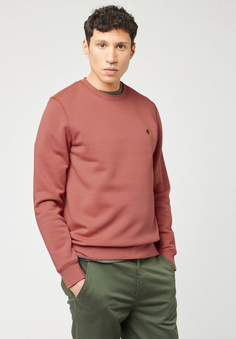 Uomo con capelli corti e ricci, che indossa un felpa color rosa polveroso e pantaloni verde oliva, con le mani nelle tasche, in piedi contro uno sfondo chiaro e semplice.