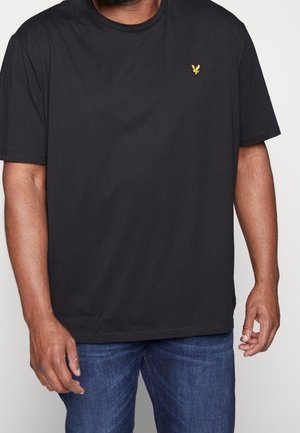 Hombre vistiendo una camiseta negra de manga corta sin estampado con un pequeño logotipo de un pájaro amarillo en el pecho izquierdo y jeans de mezclilla azul.