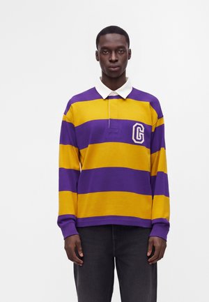 Jeune homme portant un maillot de rugby à manches longues avec larges rayures horizontales violettes et jaunes et un col blanc, debout devant un fond uni.
