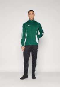 Grønn atletisk jakke med hvite striper, glidelås foran og høy krage. Sorte bukser med hvite sidestriper, sammen med svarte sko.