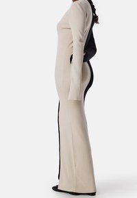 Deux personnes de dos l'une à l'autre, l'une portant une longue robe beige côtelée, l'autre en noir, toutes deux se tenant devant un fond uni.