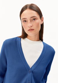 Junge Frau mit glattem braunem Haar und Sommersprossen trägt eine blaue knöpfbare Strickjacke über einem weißen Rundhals-Shirt vor einem schlichten weißen Hintergrund.