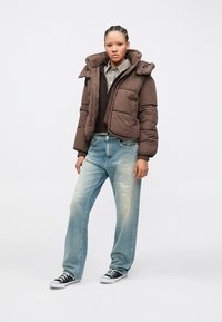 Chaqueta acolchada marrón con cuello alto, con cierre de cremallera delantero y bolsillos laterales, combinada con vaqueros azul claro desgastados y zapatillas negras.