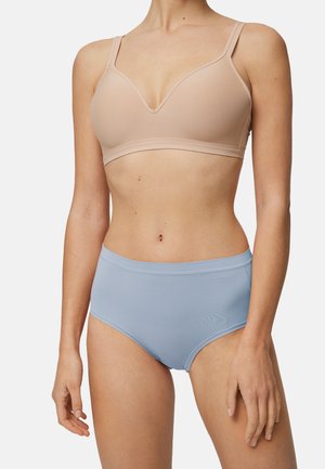 Mujer con un sujetador inalámbrico beige y bragas de tiro alto azul claro, de pie contra un fondo liso y claro.