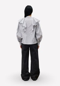 Persoon met lang donker haar, gekleed in een wit met zwart verticaal gestreept blouse met ruffles en losse zwarte broek, staand met de rug naar de kijker.