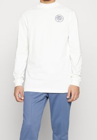 White long-sleeve shirt with a high neckline and blue "Nike Athletic Club" logo on the left chest. Smooth fabric, fitted design, minimalistic style.