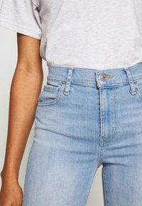 Primer plano de una persona que lleva jeans de talle alto de color azul claro y una camiseta de color gris claro metida por dentro, mostrando la cintura y la parte superior del muslo.