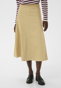 Jupe beige à mi-mollet au design évasé, avec des coutures visibles et une texture lisse. Associée à des mocassins noirs.