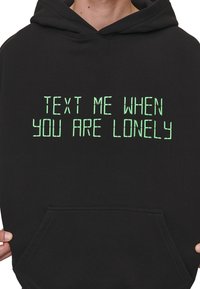 Black hoodie with a front pocket and green graphic text reading "TEXT ME WHEN YOU ARE LONELY." Cotton material with a soft texture.