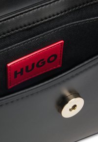 Sort lædertaske med en tekstureret inderside, der har en fremtrædende rød HUGO-tag og guldfarvet hardware på lukningen.