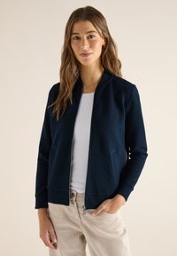Femme aux longs cheveux bruns portant une veste zippée bleu marine sur un haut blanc et un pantalon beige, debout avec une main dans la poche sur un fond neutre.