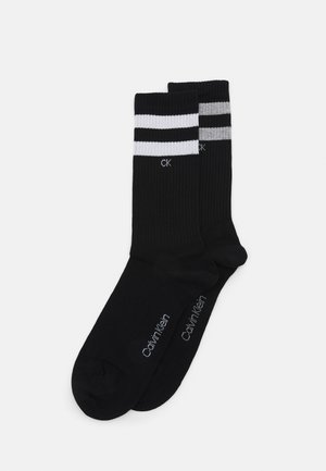 Calcetines de algodón negros con rayas blancas y grises en la parte superior. Tienen una textura acanalada y el logo "Calvin Klein" impreso cerca de la zona del dedo del pie.