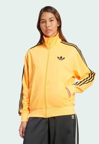 Gelbe Zip-Jacke für das Training mit schwarzen Streifen an den Ärmeln, mit einem kleinen schwarzen Logo auf der Brust und seitlichen Taschen; glatte Textur.