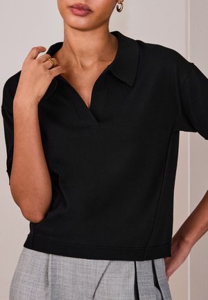 Ženska v črni kratkostični majici s ovratnikom in V-izrezom ter sivih nabrkanih hlačah, ki se dotika ogrlice, vidni so zlati obročasti uhani in prstan.