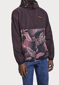 Man med mörklila huva jacka med rosa tropiskt bladmönster framtill, ihop med blå jeans, stående mot en enhetlig bakgrund.