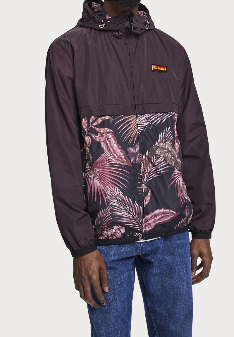 Man med mörklila huva jacka med rosa tropiskt bladmönster framtill, ihop med blå jeans, stående mot en enhetlig bakgrund.