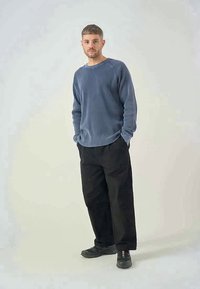 Charcoal long-sleeve waffle knit shirt paired with loose black trousers and dark shoes, standing against a light background.