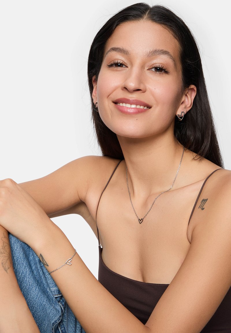 Gioielli color argento con dettagli a forma di cuore, abbinati a una maglietta marrone e jeans. Il tono della pelle è caldo e i capelli sono lunghi e scuri.