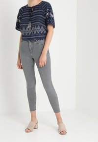 Top corto con patrón de marina y mangas cortas, combinado con jeans skinny grises y sandalias de tiras beige. Presenta un lazo con borlas en el escote.