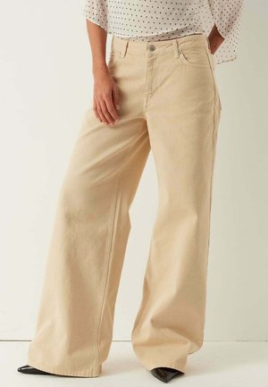 Beige wijde broek gedragen met een witte blouse met zwarte stippen, gecombineerd met zwarte puntige schoenen, getoond van taille tot voeten.
