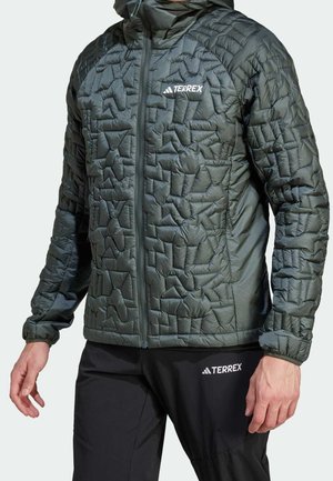 Uomo che indossa una giacca nera trapuntata Adidas Terrex e pantaloni neri Terrex, in piedi con le braccia rilassate lungo i fianchi.