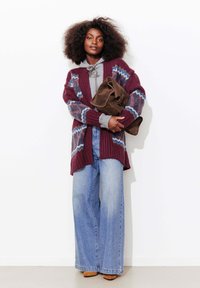 Cardigan en maille bordeaux avec motifs bleus et blancs, sweat à capuche gris en dessous, assorti à un jean large bleu clair et un sac marron.