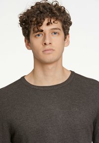 Brun stickad tröja med rund halsringning. Har en mjuk textur och subtil logga vid bröstet. En enkel design utan mönster eller detaljer.