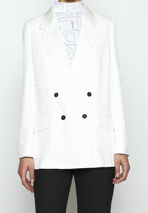 Blazer blanc croisé avec un motif de texte embossé subtil, boutons noirs, porté sur un top blanc à col montant avec motif de texte noir.