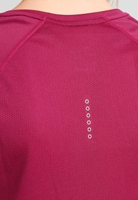 Camiseta deportiva fucsia con textura de malla, que presenta un cuello redondo y seis pequeños detalles metálicos a lo largo de la parte superior de la espalda.