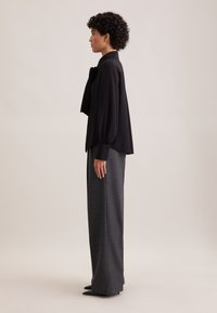Schwarze Bluse mit langen Ärmeln und einer Schleifendetail am Kragen, kombiniert mit hochgeschnittenen grauen Hosen. Glatte Materialien mit einer lockeren Passform.