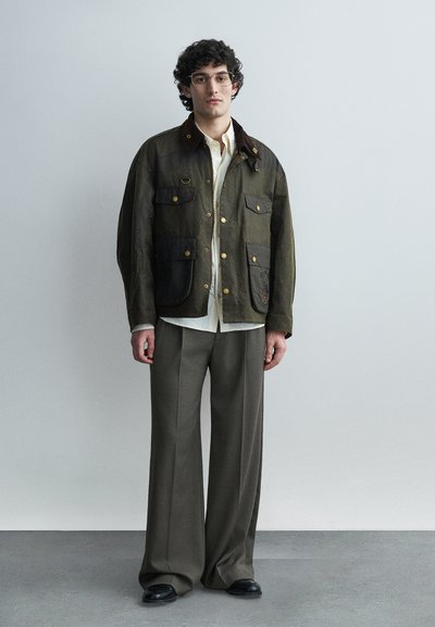 Νεαρός άνδρας με γυαλιά, φορεμένο ένα χακί μπουφάν με πολλές τσέπες, λευκό πουκάμισο, γκρι παντελόνι φαρδύ και μαύρα παπούτσια, στέκεται μπροστά σε απλό φόντο.