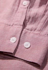 Dettaglio di una camicia di lino rosa chiaro con colletto piegato e due bottoni bianchi in plastica, che mette in evidenza un tessuto testurizzato e cuciture pulite.