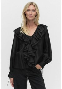 Svart blus med volangkrage, långa ärmar och samlade manschetter; slät textur och avslappnad passform; matchas med mörka jeans med dekorationer.