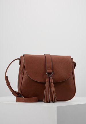 Bruine leren crossbodytas met klep en twee kwastjes, verstelbare schouderriem, rustend op een wit voetstuk.