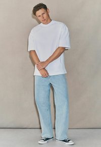 Camiseta blanca de gran tamaño y manga corta, pantalones de mezclilla ligeros en azul, y zapatillas de lona negras con suelas blancas. Texturas suaves, ajuste relajado.