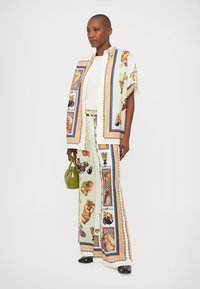 Costum format din două piese, cu o cămașă patternată și pantaloni largi, dintr-un material ușor cu ilustrații de fructe și vaze. Accentuat cu o geantă verde.