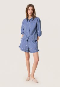Camicia di cotone blu con motivi ricamati bianchi, abbinata a pantaloni corti coordinati con orli arricciati. Scarpe piatte semplici completano l'outfit.