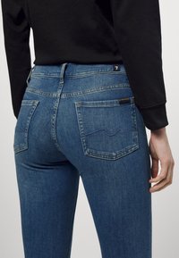 Blue denim jeans with a fitted design, featuring two back pockets, subtle stitching detail, and a visible tag on the waistband.