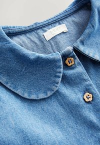 La chemise en denim présente une couleur bleu clair, un col arrondi et deux boutons décoratifs en bois en forme de fleur au niveau du col. Étiquette de marque visible.