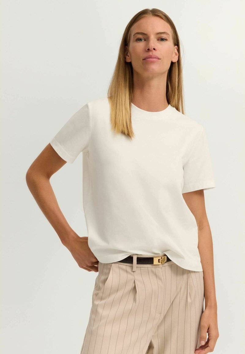 Frau mit glatten blonden Haaren, die ein schlichtes weißes T-Shirt und beige gestreifte Hosen mit einem schwarzen Gürtel trägt, steht mit einer Hand in der Hüfte.