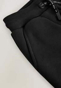 Mustad vöökohaga sportpüksid elastse vöökohaga ja nähtava küljetaskuga, millel on must tõmbelint valgete kirjadega.