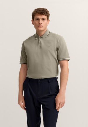 Khaki polo shirt with a collar, three buttons, and contrasting striped trim on sleeves, paired with tailored navy trousers.