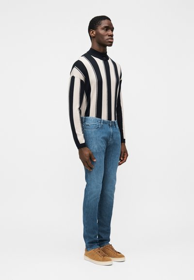 Pleteni črni in bež pulover z visokim ovratnikom z črtami, modre jeans hlače iz jeansa, rjavi semiš športni copati, prilegajoč se kroj, gladka tekstura, srednje modra obdelava.