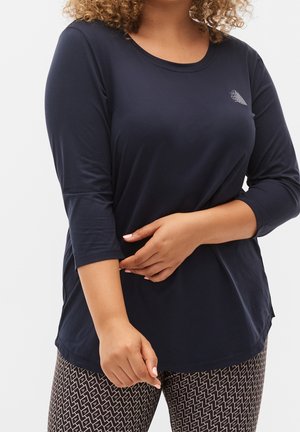 Femme portant un t-shirt à manches longues bleu marine avec un petit logo sur la poitrine et un legging noir et blanc à motifs, les bras croisés.