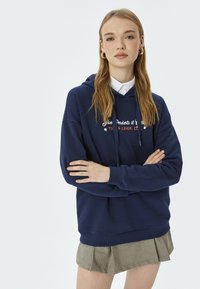Granatowy hoodie z białym i czerwonym napisem, z kapturem ze sznurkiem, ściągaczami i luźnym krojem, w połączeniu z plisowaną spódnicą.