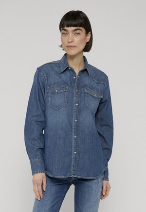 Donna con capelli corti e scuri indossa una camicia di jeans abbottonata con tasche con patta e jeans blu su uno sfondo grigio semplice.