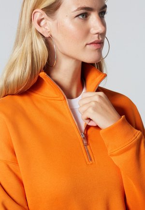 Oranger Sweatshirt mit Reißverschluss, hohem Kragen, gerippten Bündchen und glatter Baumwollstruktur, kombiniert mit einem weißen Hemd darunter.